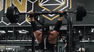 Strongman Evan Singleton in gym pressing heavy barbell overhead.