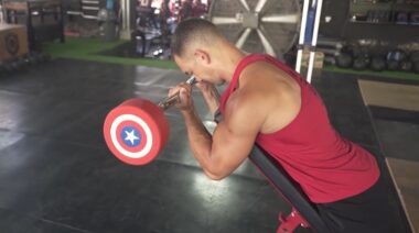 muscular person in gym leaning on bench doing barbell curl